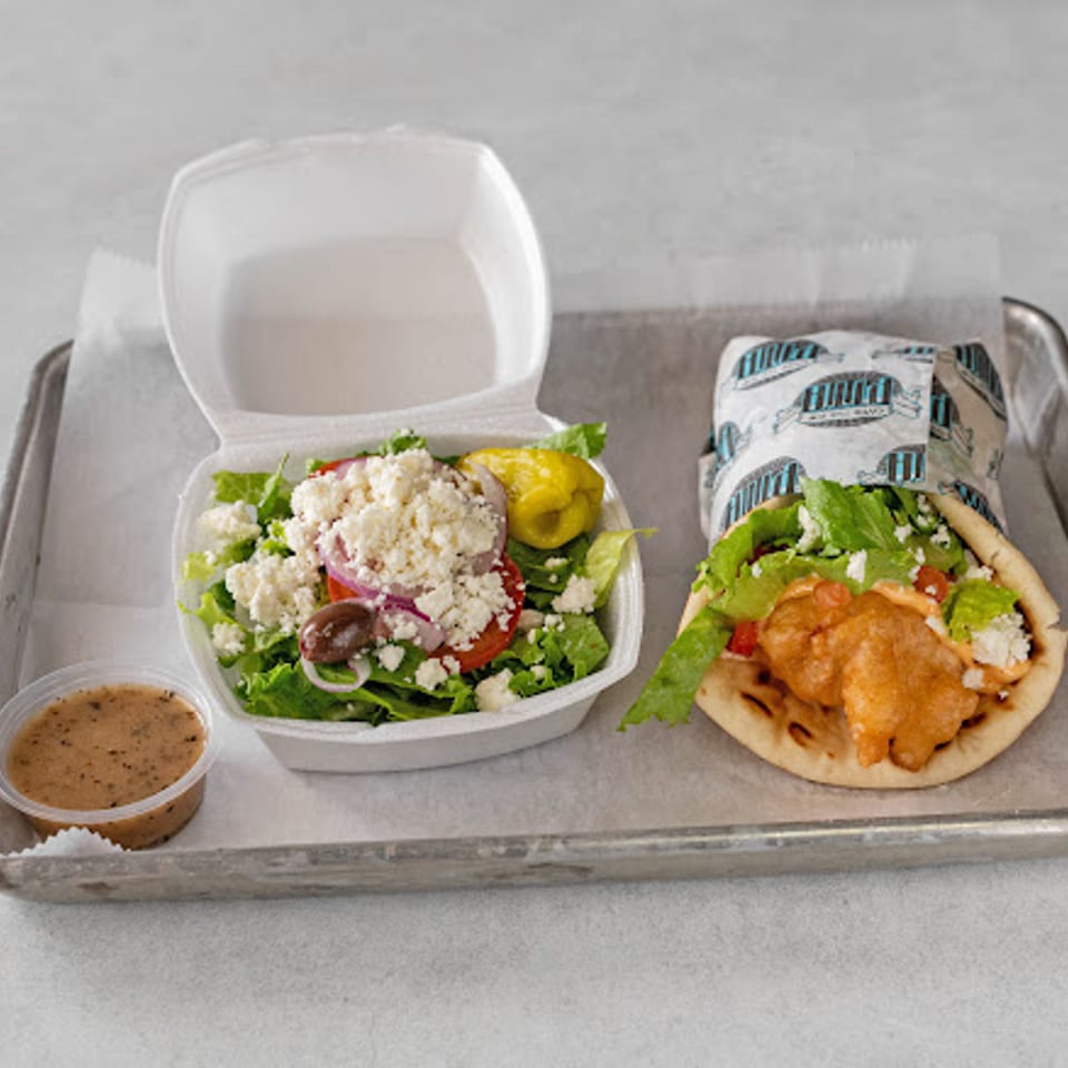 Tray with Greek salad, pita sandwich filled with crispy chicken and lettuce, wrapped flatbread, and dipping sauce.
